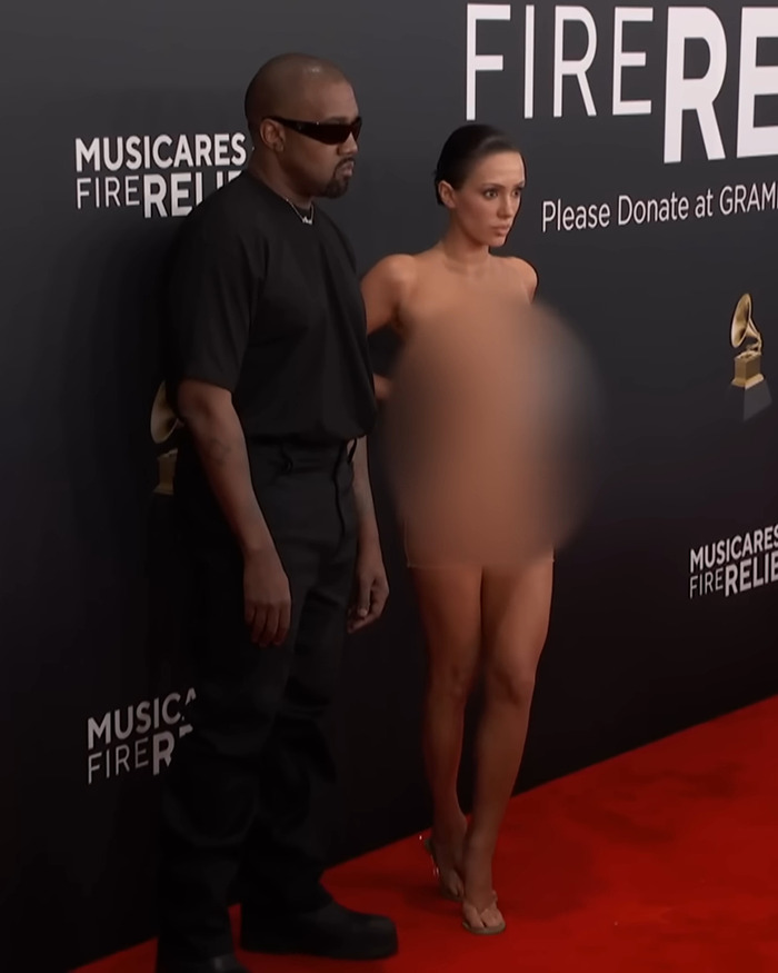Kanye West in black attire and sunglasses with Bianca Censori in blurred outfit on Grammys red carpet.