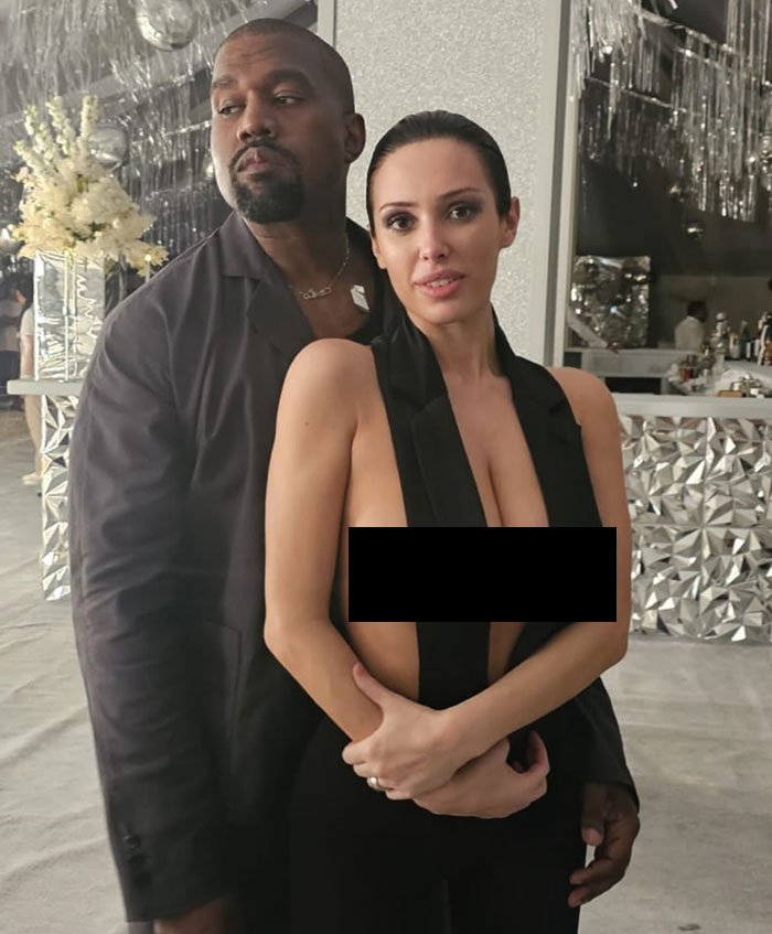 A person posing in a revealing black outfit, standing with a man in a decorative room, featuring Bianca Censori. A person posing in a revealing black outfit, standing with a man in a decorative room, featuring Bianca Censori.