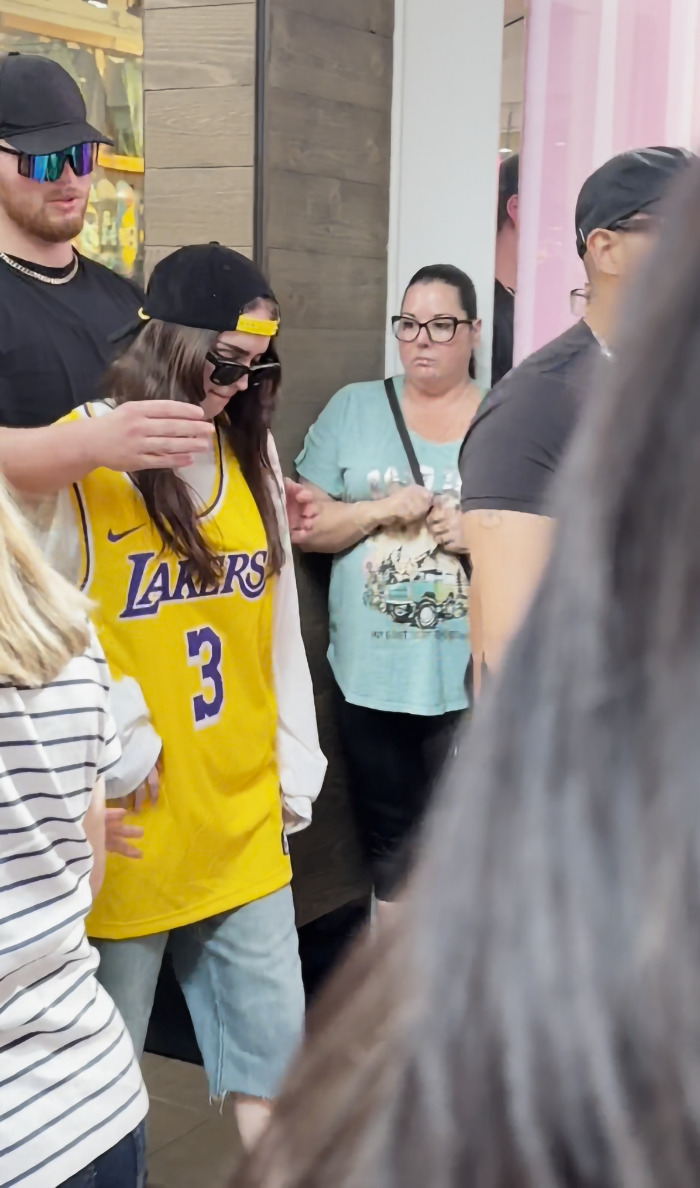 Billie Eilish look-alike in yellow Lakers jersey surrounded by people at a busy mall event.