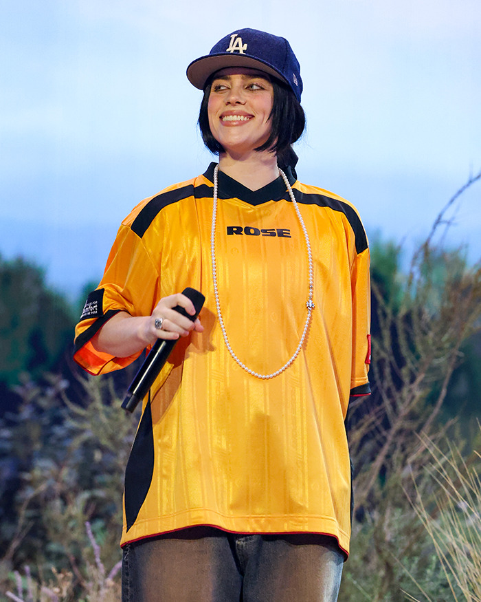 A person resembling Billie Eilish in a yellow jersey and cap, holding a microphone outdoors.