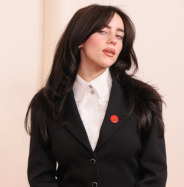 Billie Eilish in a formal outfit with a red pin, posing for a portrait.