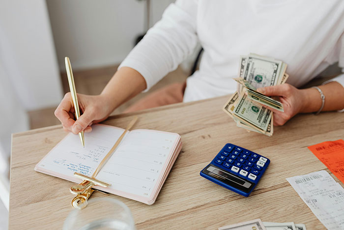 A woman managing finances at home, writing in a notebook, counting cash, with a blue calculator nearby. A woman managing finances at home, writing in a notebook, counting cash, with a blue calculator nearby.