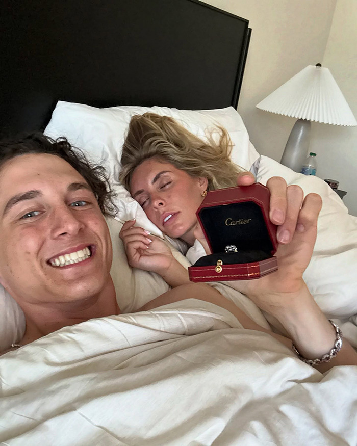 A man smiling while holding a Cartier ring box next to a sleeping woman in bed.