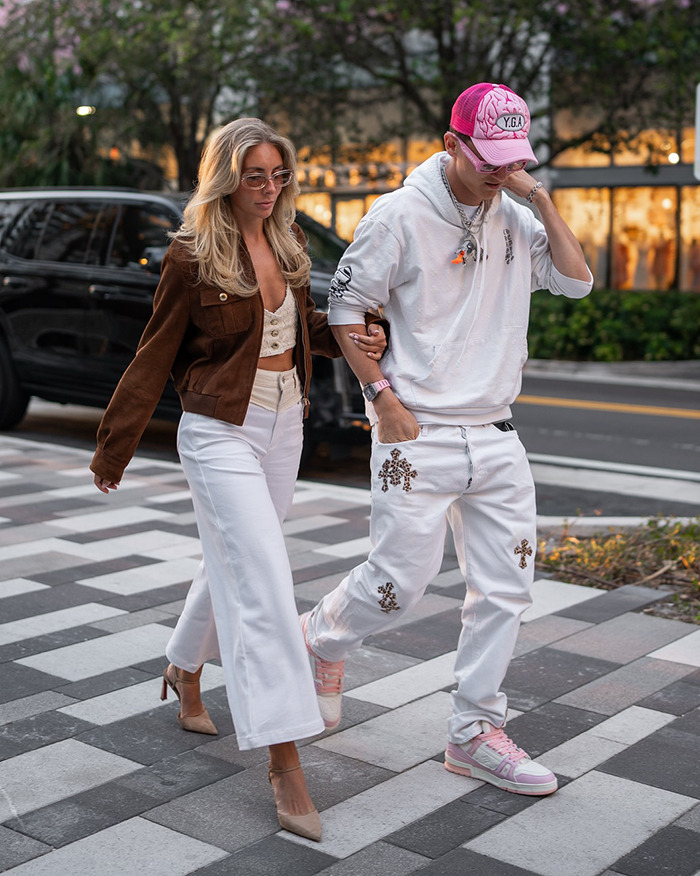 Couple walking on a sidewalk; woman wears sunglasses and brown jacket, man in pink cap and white outfit with patterns.