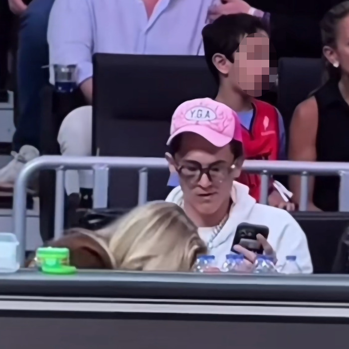 Person at NBA event causing outrage, wearing glasses and a pink cap, seated and looking at phone with audience in background.