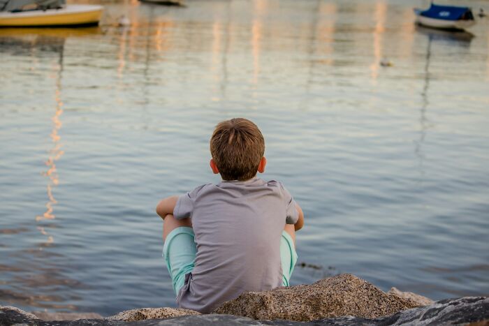 Boy in a gray shirt and green shorts sitting on rocks by the water, reflecting peacefully.
