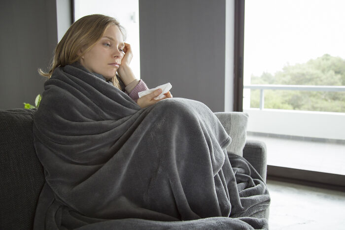 Woman wrapped in a gray blanket, sitting on a sofa, looking out a window, appearing thoughtful and relaxed.