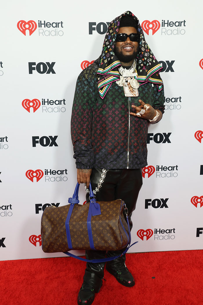 A person in a multicolored outfit with a large bag at the 2025 iHeartRadio Music Awards red carpet.