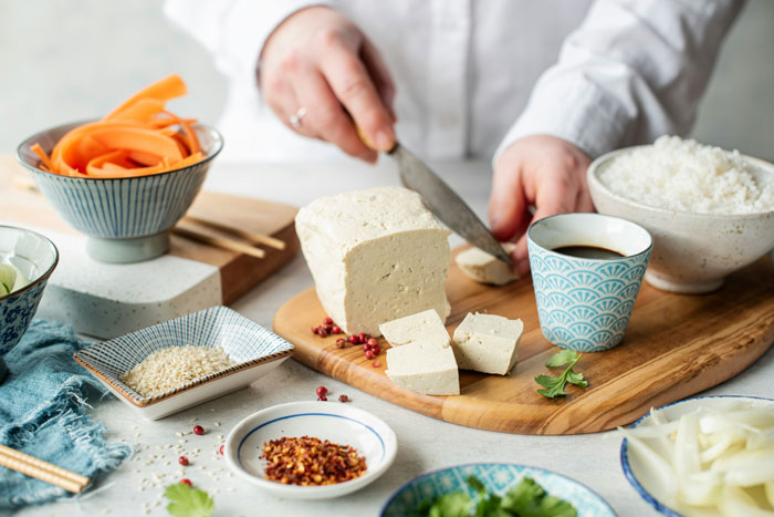 Person slicing tofu on a wooden board, surrounded by vegetables, a bowl of rice, and various spices, related to cooking.