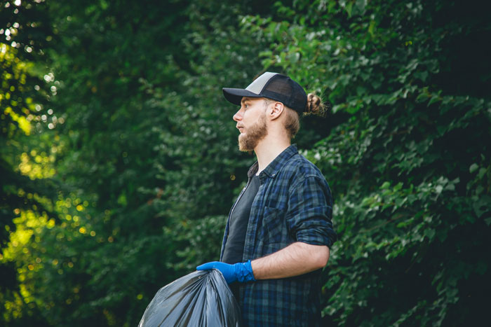 Man outdoors holding a trash bag, wearing a cap and checkered shirt, representing a boyfriend discarding sentimental items. Man outdoors holding a trash bag, wearing a cap and checkered shirt, representing a boyfriend discarding sentimental items.