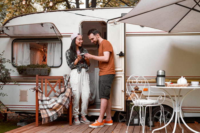 Couple smiling outside a camper, with a table set for coffee; surrounded by trees and nature. Couple smiling outside a camper, with a table set for coffee; surrounded by trees and nature.