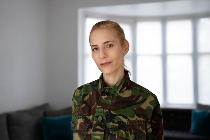 Woman in a camo jacket stands in a living room, expressing emotions related to motherhood fears.