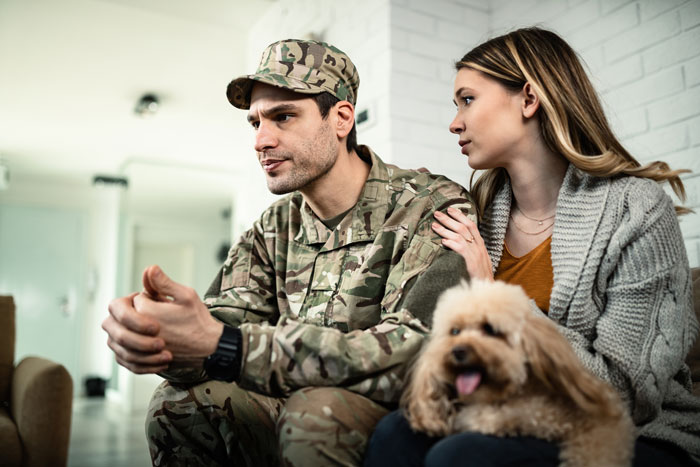 Couple discussing motherhood, man in military uniform, woman concerned, comforting dog nearby.