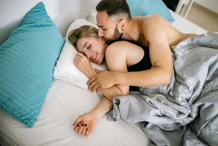 Couple cuddling in bed, both smiling, with gray blankets and blue pillows, highlighting open relationship dynamics.