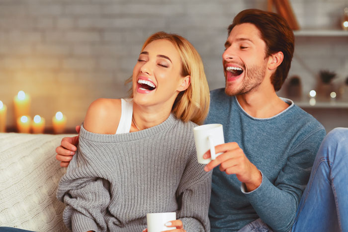 A couple laughing on a sofa, each holding a mug, in a cozy living room setting.