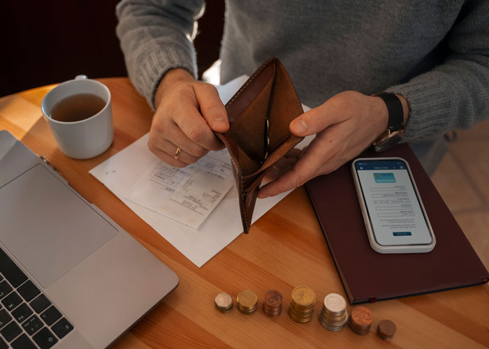 A man opens an empty wallet, surrounded by coins and bills, illustrating financial struggles and spending habits.