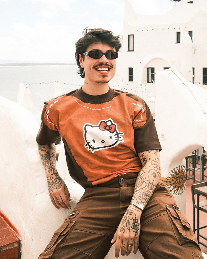 Tattooed person in sunglasses and Hello Kitty shirt, sitting outdoors with Brazilian architecture in background.