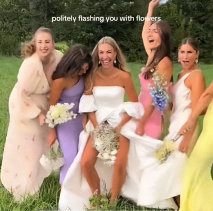 Bride flashes underwear during wedding photoshoot with bridesmaids holding flowers, laughing in a grassy field.