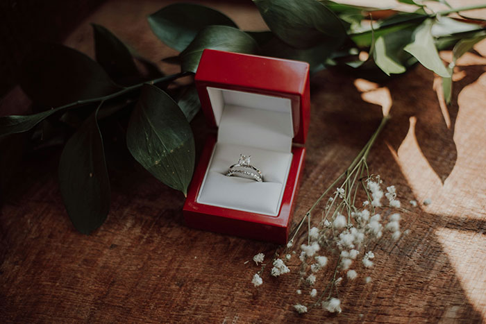 Red ring box on a wooden table with greenery, highlighting an engagement ring and wedding themes.