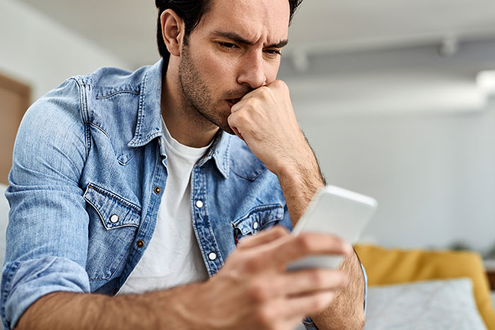 Man in denim shirt looking at phone, appearing thoughtful while considering engagement at a friend's wedding.
