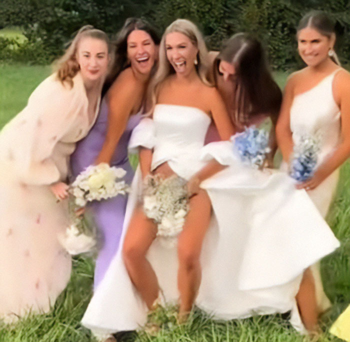 Bride flashes underwear with bridesmaids during wedding photoshoot in a playful outdoor setting.
