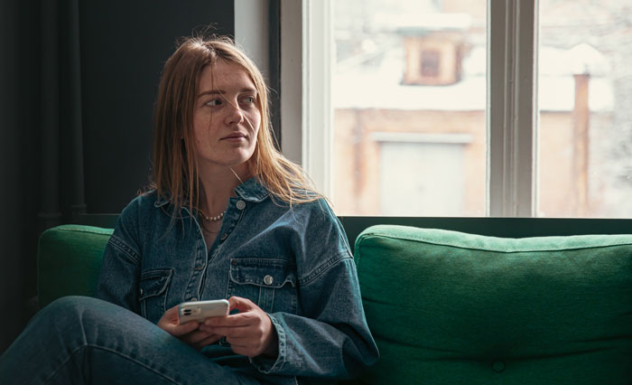 Woman in denim on green sofa, holding a phone, looking thoughtful about immigrants.