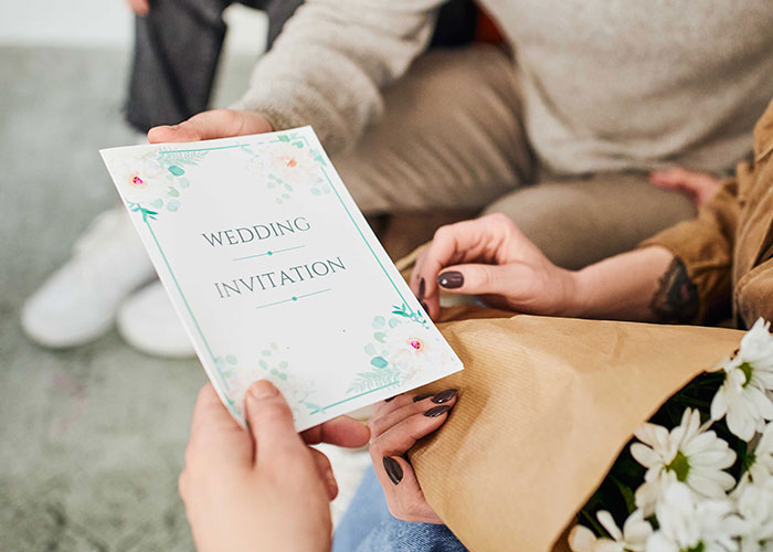 People holding a wedding invitation card, surrounded by flowers and casual attire, suggesting a discussion about wedding plans.