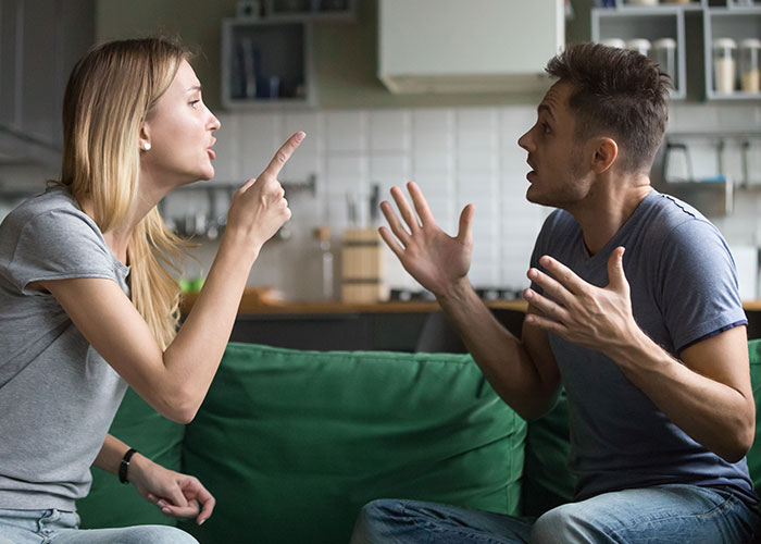Man and woman arguing on the couch, emphasizing relationship tension with his choice of plus-one to the wedding.