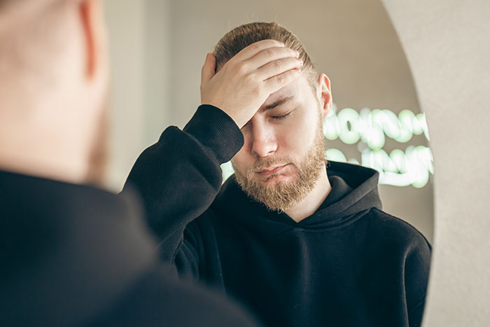 Man in black hoodie looking distressed, hand on forehead, in front of a mirror.