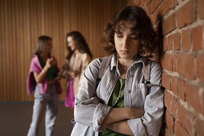 Teen bullied over ridiculous things, leaning against a brick wall while two girls whisper.