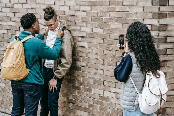 A student intimidates another against a brick wall while a third films with a phone, highlighting bullying issues.