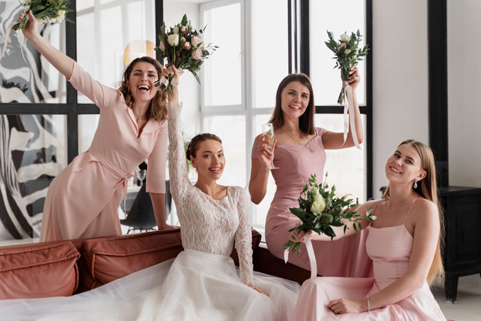 Bridesmaids in pink dresses and bride smiling and holding bouquets indoors, with natural light.