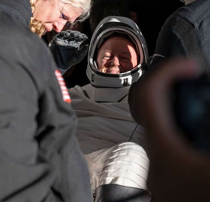NASA astronaut in suit, smiling after Earth return, with concern for his health being addressed by the crew.