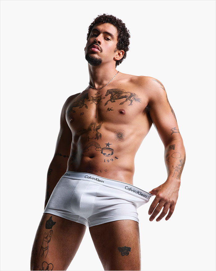 Man posing shirtless in a Calvin Klein ad, showcasing tattoos and white briefs.