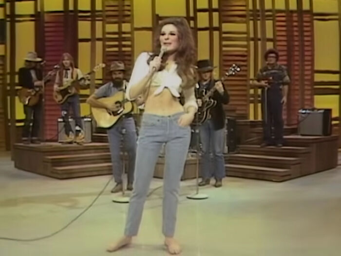 Singer performing barefoot on stage with a band, wearing jeans and a tied shirt. Celebs who disappeared from the media.