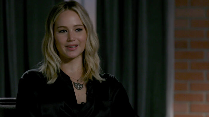 A celebrity in a dark outfit, smiling, seated indoors with a brick wall background, keeping it classy.