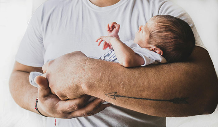 Man in white shirt holding a newborn, with a tattooed arm.