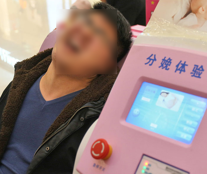 A man undergoing childbirth simulation, seated in front of a pink medical device. A man undergoing childbirth simulation, seated in front of a pink medical device.