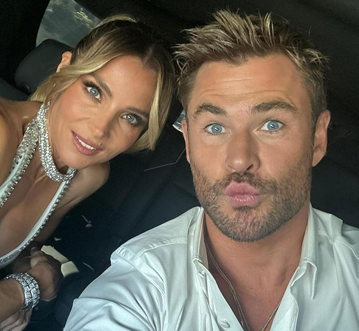 Chris Hemsworth taking a playful selfie in a car, wearing a white shirt, alongside another person.