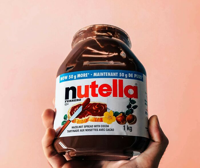 A hand holding a 1kg jar of Nutella against a peach background; some find it unappealing despite its popularity.