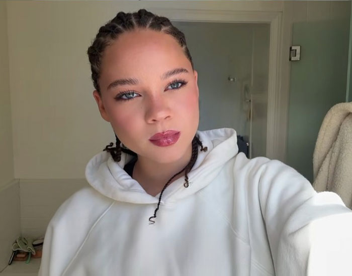 Young woman in a white hoodie, braided hair, posing confidently in a bright room.