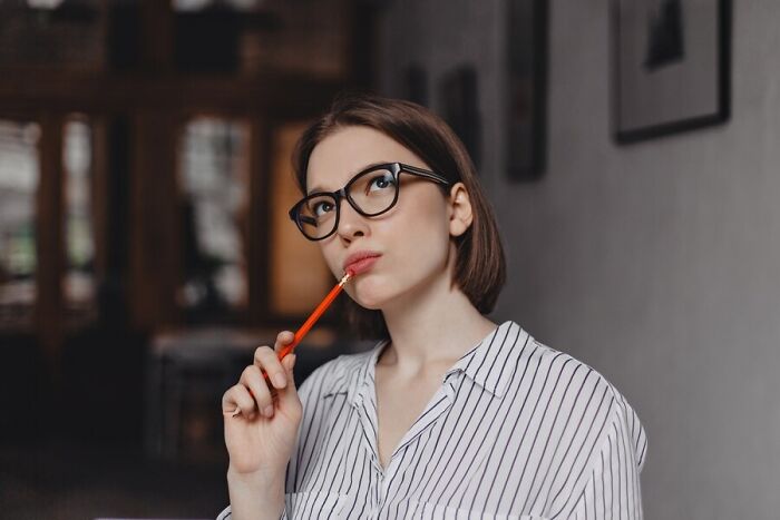 Woman in striped shirt with glasses, pondering with pen in mouth, illustrating signs of being subtly smarter.