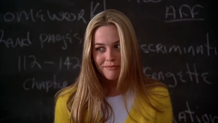 Blonde woman in yellow cardigan with blackboard background, evoking Clueless nostalgia.