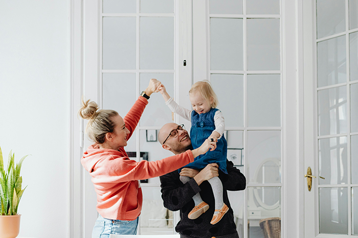 Family moment in a bright room, with parents lifting their child playfully near a large window. Family moment in a bright room, with parents lifting their child playfully near a large window.