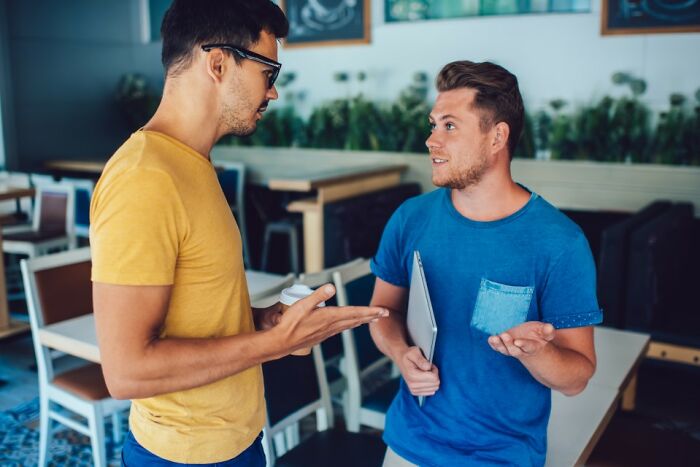 Two men in a cafe having an intense discussion about former best friends, one holding a laptop.