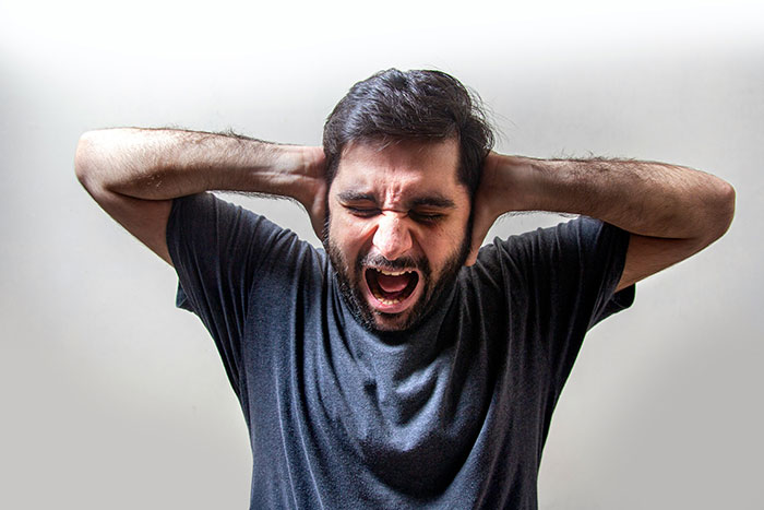Man in gray shirt covering ears, appearing distressed, illustrating psychological tricks and emotional reaction.