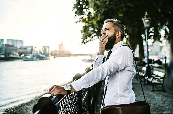 A man in a white shirt yawning on a riverside walkway, demonstrating a psychological trick in a relaxed setting.
