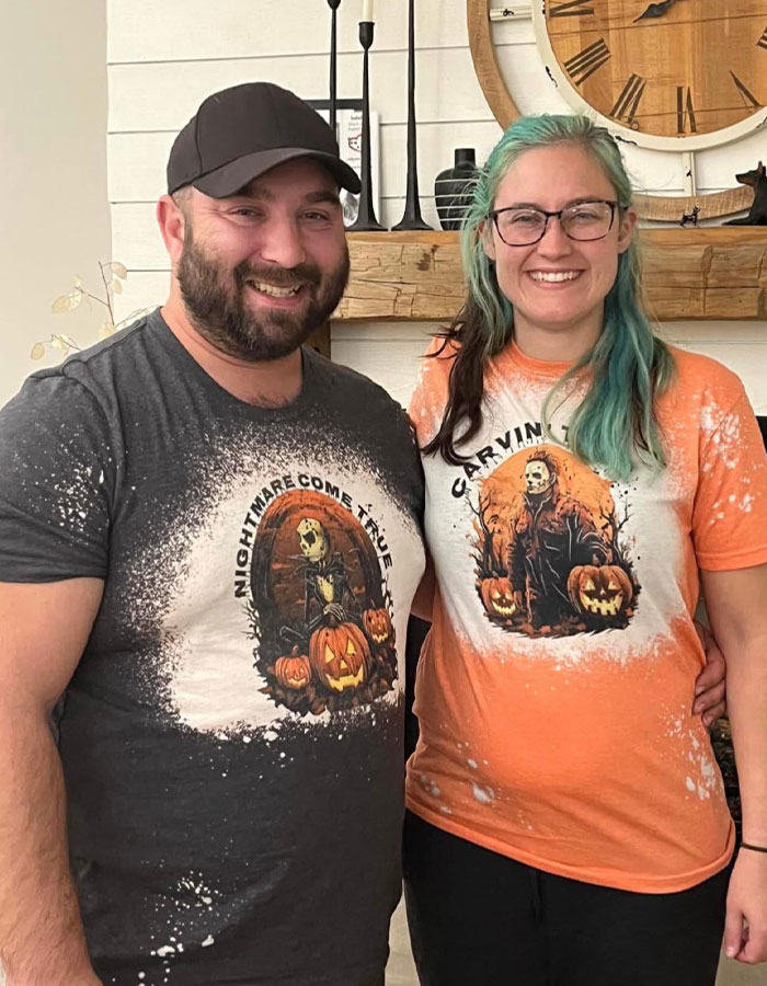 A smiling couple wearing Halloween-themed shirts with pumpkin designs, standing indoors.