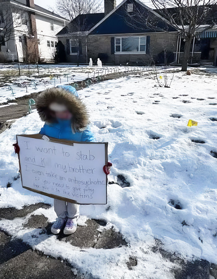 Young child in winter coat stands in snow holding a sign as a police officer's punishment.