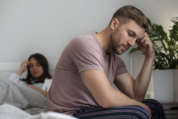 Man sitting on bed, appearing thoughtful with hand on forehead, woman in the background.